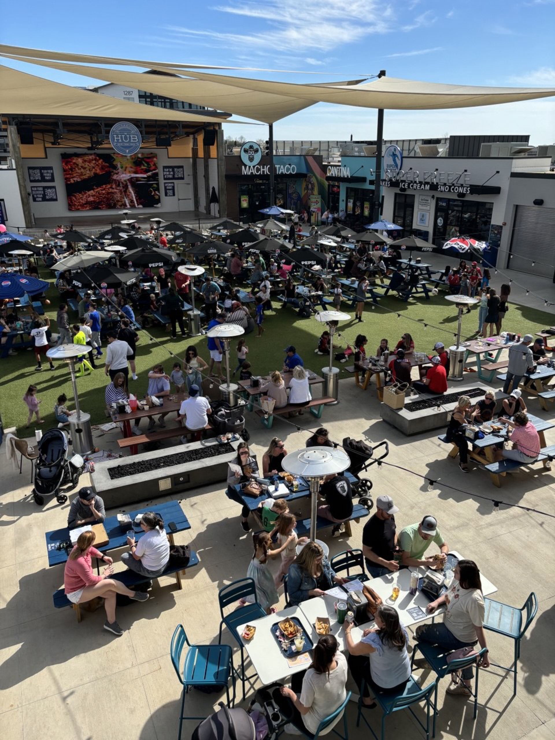 Packed daytime courtyard with families, restaurants, and Jumbotron at The HUB